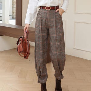 以下が含まれることがあります： 白いボタンダウンシャツ、茶色のチェック柄パンツ、茶色の革ブーツを組み合わせた女性の服装。 金色のバックルが付いた茶色の革ベルトと茶色の革のハンドバッグがアンサンブルを完成させます。 パンツはハイウエストでゆったりとしたフィット感です。