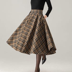 Puede incluir: Una mujer con un top negro de manga larga y una falda a cuadros marrón. La falda tiene un diseño acampanado y presenta un patrón de cuadros clásico en tonos marrón, negro y blanco. También lleva medias negras y tacones negros.