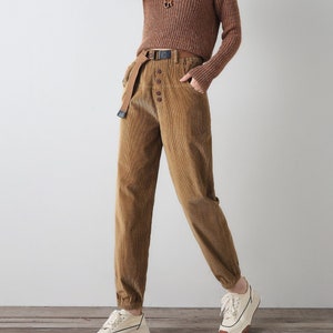 K&ouml;nnte beinhalten: Ein brauner Rollkragenpullover mit einer braunen Cordhose und wei&szlig;en Turnschuhen. Die Hose hat einen braunen G&uuml;rtel und Knopfdetails.
