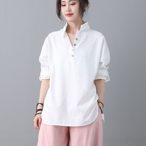 May include: A white, long-sleeved, collared shirt with a single button closure at the neckline. The shirt has a relaxed fit and a slightly curved hemline. The model is wearing the shirt with pink pants.