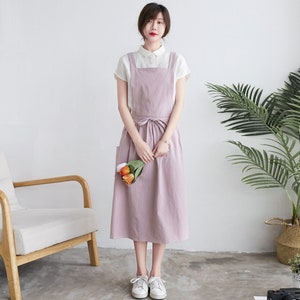 May include: A light pink linen apron dress with a white top and a tie at the waist. The dress has two pockets on the front.