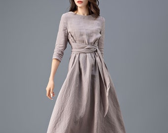 May include: A woman wearing a long, light brown linen dress with a self-tie belt. The dress has a round neckline and three-quarter sleeves.