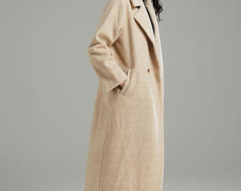 May include: A long, beige wool coat with a classic lapel collar and two front pockets. The coat is worn over a black top and blue jeans. The model is wearing black ankle boots with silver chain detailing.