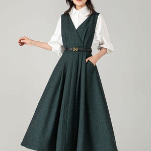 May include: A dark green dress with a V-neck design and a flared skirt. The dress is paired with a white blouse and a black belt with a gold buckle. The outfit is completed with black heels.
