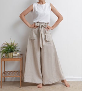 May include: A woman wearing a white sleeveless top and wide-legged linen pants with a self-tie belt. The pants are a light beige color and have pockets on the sides.