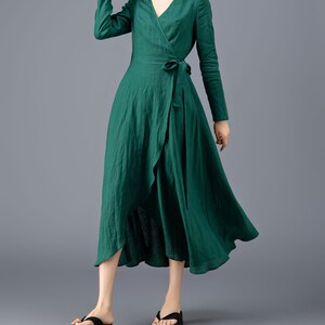 May include: A green wrap dress with long sleeves and a tie at the waist. The dress has a high neckline and a flowing skirt. The model is wearing a straw hat and black sandals.