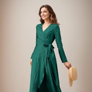 May include: A woman wearing a green wrap dress with long sleeves and a tie at the waist. She is holding a straw hat in her right hand and walking towards the camera.