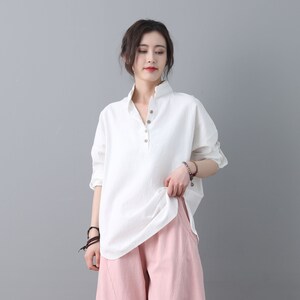 May include: A white linen shirt with a button-up collar and long sleeves. The shirt has a relaxed fit and is slightly oversized. The model is wearing the shirt with pink pants.