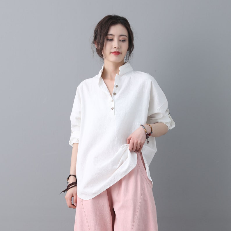May include: A white linen shirt with a button-up collar and long sleeves. The shirt has a relaxed fit and is slightly oversized. The model is wearing the shirt with pink pants.