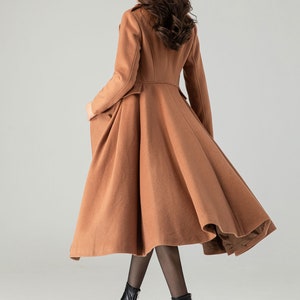 May include: A long, brown, wool coat with a flared skirt and a fitted bodice. The coat has a high collar and long sleeves. The model is wearing black tights and black heeled boots.