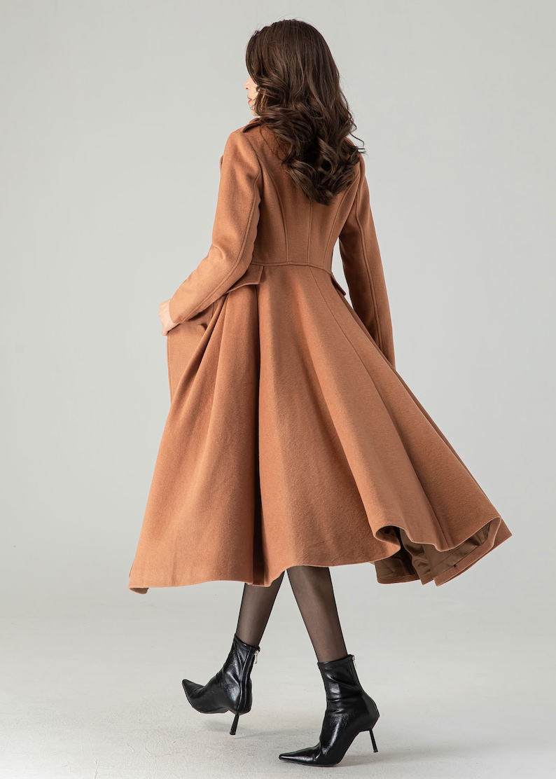 May include: A long, brown, wool coat with a flared skirt and a fitted bodice. The coat has a high collar and long sleeves. The model is wearing black tights and black heeled boots.