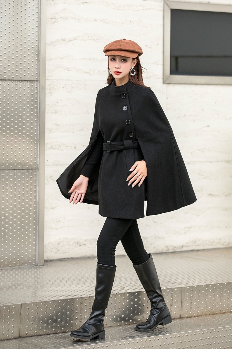 May include: A black wool cape with a belt and buttons. The cape has a wide, flowing design and is worn over a black turtleneck and black pants. The person is wearing black boots and a brown newsboy cap.