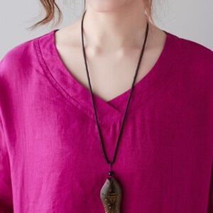 May include: A magenta V-neck shirt with long sleeves. A black cord necklace with a wooden fish pendant hangs from the neckline. The shirt is made of a soft, flowing fabric.
