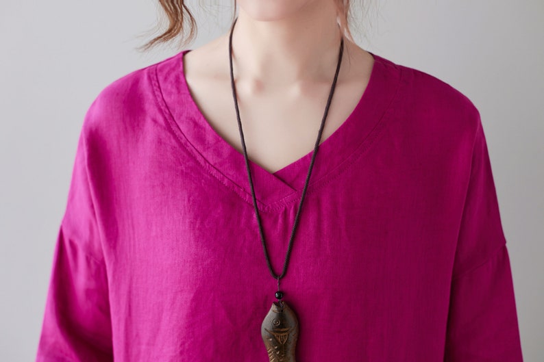May include: A magenta V-neck shirt with long sleeves. A black cord necklace with a wooden fish pendant hangs from the neckline. The shirt is made of a soft, flowing fabric.