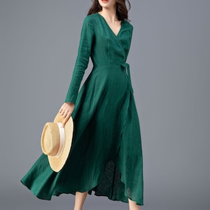 May include: A woman wearing a long-sleeved, emerald green wrap dress with a tie at the waist. She is holding a straw hat in her hand and is standing on a gray background.