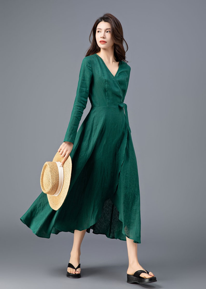 May include: A woman wearing a long-sleeved, emerald green wrap dress with a tie at the waist. She is holding a straw hat in her hand and is standing on a gray background.