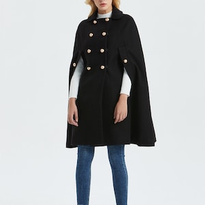 May include: A black double-breasted cape with gold buttons and a long, flowing silhouette. The cape is worn over a white turtleneck sweater and blue jeans.