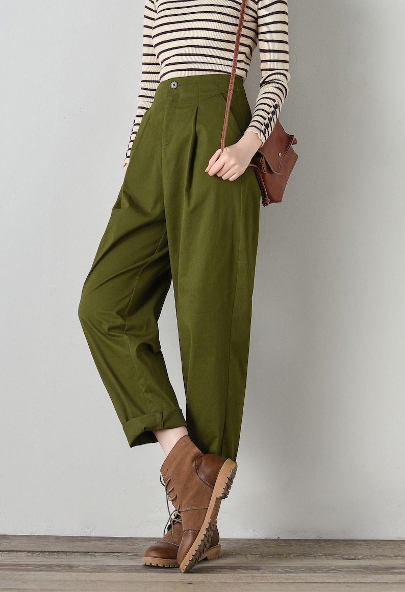 Op de afbeelding: Een olijfgroene broek met hoge taille, een knoopsluiting en plooien. De broek is aan de onderkant gezoomd en het model draagt ​​bruine leren laarzen.