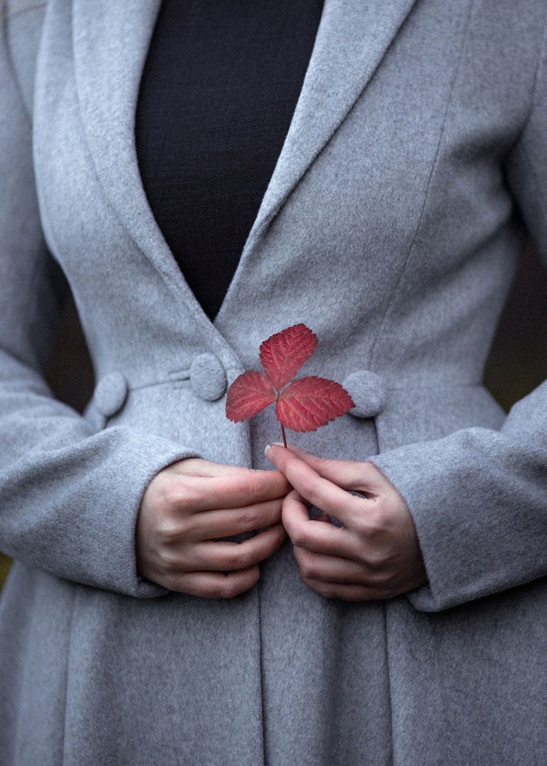 May include: A light gray coat with a black top underneath. A red leaf is held in front of the coat. The coat has three buttons and a classic design. The image has a soft focus.