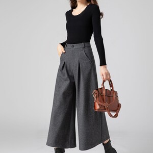 Pu&ograve; includere: Una donna indossa una camicia nera a maniche lunghe e pantaloni larghi grigi. Porta una borsa a mano in pelle marrone con una tracolla a catena dorata.