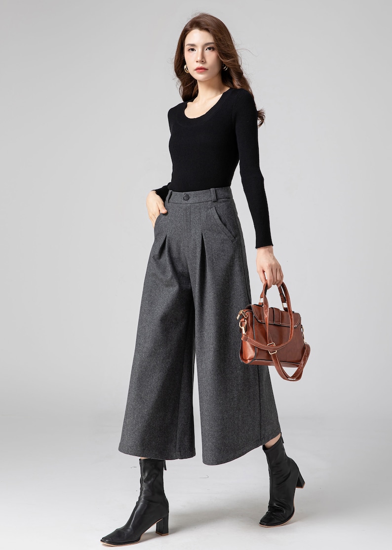 Pu&ograve; includere: Una donna indossa una camicia nera a maniche lunghe e pantaloni larghi grigi. Porta una borsa a mano in pelle marrone con una tracolla a catena dorata.