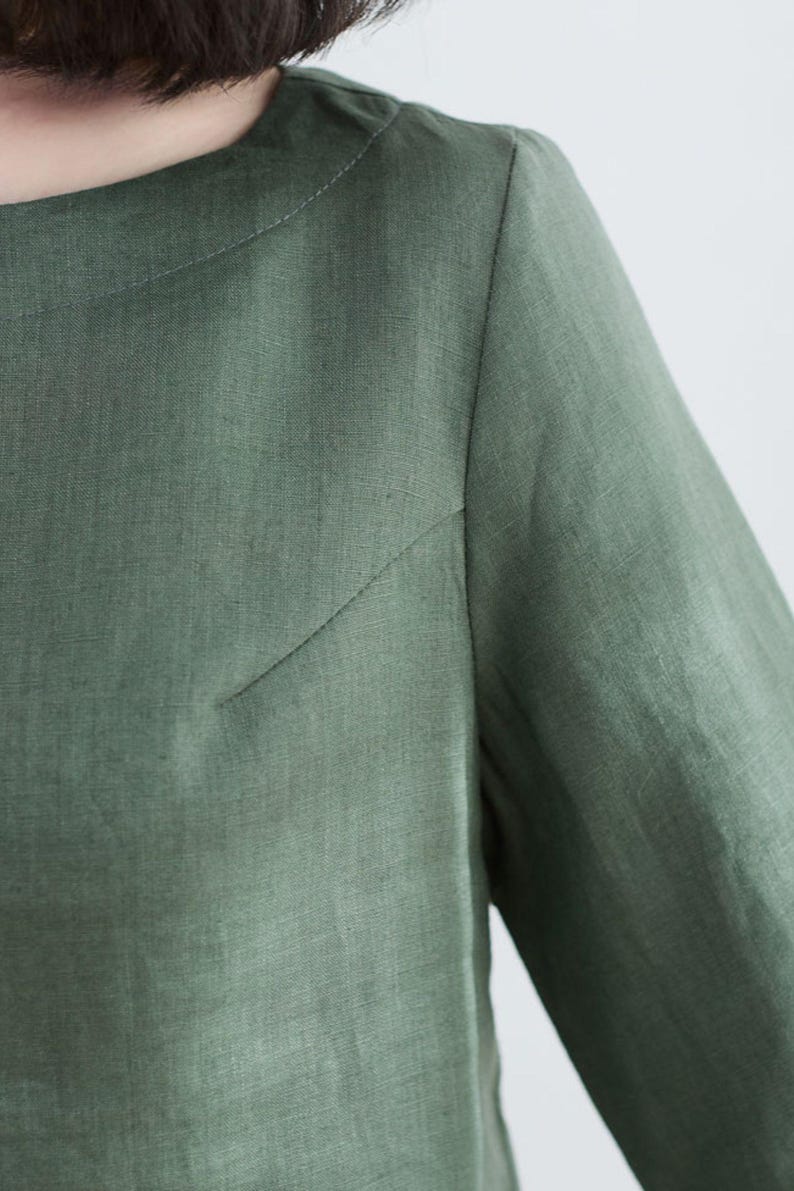 May include: A close-up of a green linen shirt with a round neckline and long sleeves.