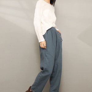 May include: A woman wearing a white long-sleeved shirt and blue linen pants with a button detail. She is wearing brown lace-up boots.