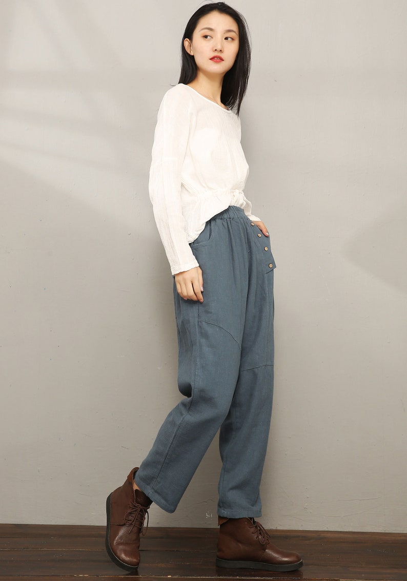 May include: A woman wearing a white long-sleeved shirt and blue linen pants with a button detail. She is wearing brown lace-up boots.
