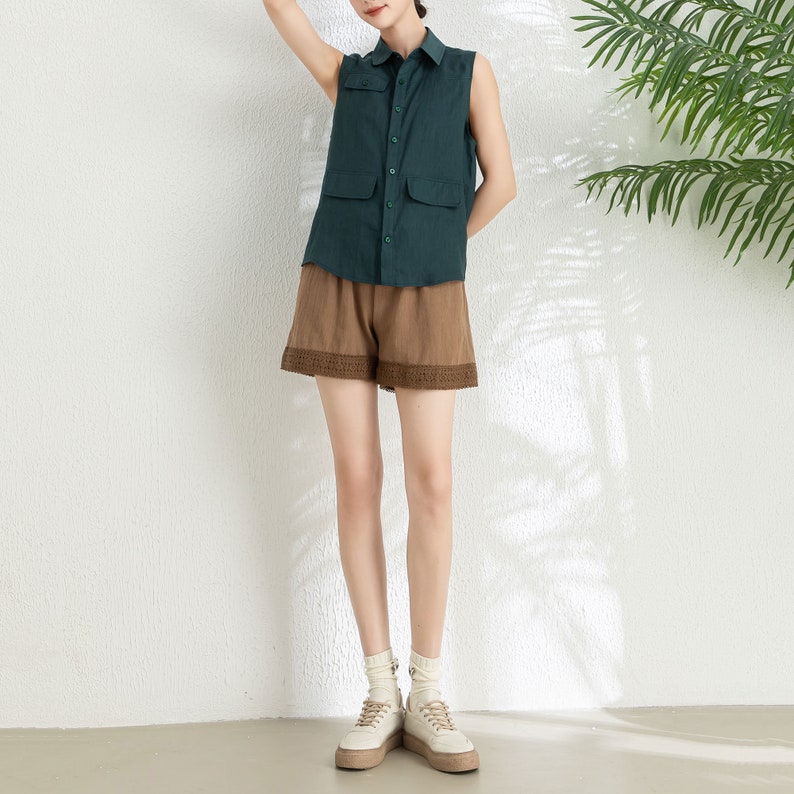 May include: A sleeveless green linen shirt with a button-up front and a brown linen skirt with lace trim. The model is wearing white sneakers.