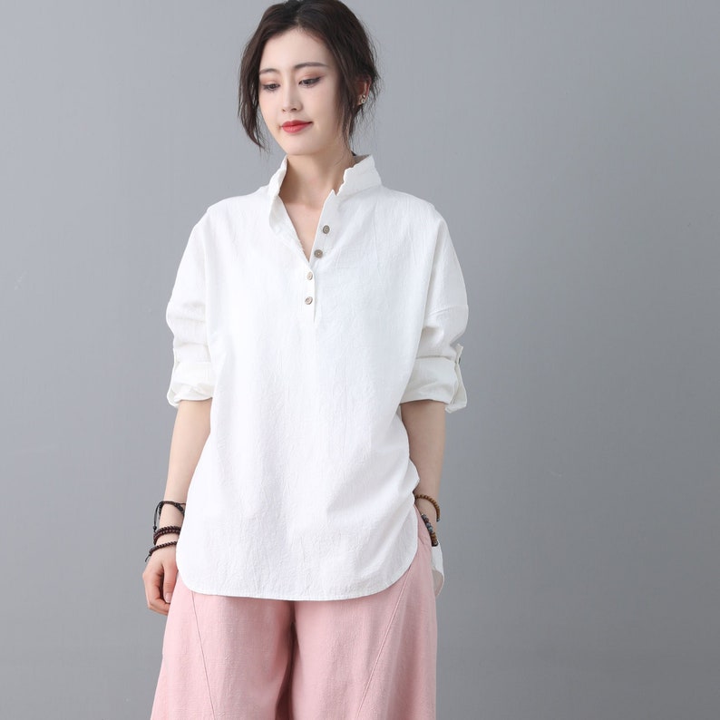 May include: A white, long-sleeved, collared shirt with a single button closure at the neckline. The shirt has a relaxed fit and a slightly curved hemline. The model is wearing the shirt with pink pants.