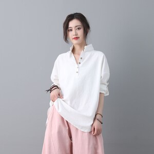 May include: A white, long-sleeved shirt with a button-up collar and a single button closure. The shirt is loose-fitting and has a relaxed silhouette. The model is wearing the shirt with pink pants.