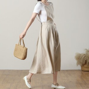 May include: A beige linen overall dress with a white t-shirt underneath. The dress has a wide, flowing skirt and adjustable straps. The model is wearing white flats and carrying a woven straw tote bag.