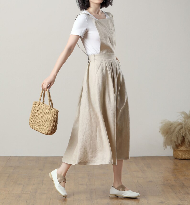 May include: A beige linen overall dress with a white t-shirt underneath. The dress has a wide, flowing skirt and adjustable straps. The model is wearing white flats and carrying a woven straw tote bag.