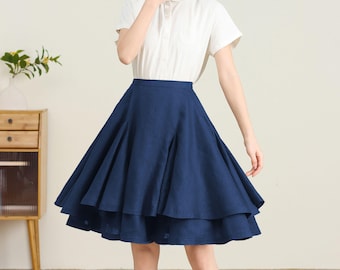 May include: A navy blue, layered skirt with a flared silhouette. The skirt is paired with a white short-sleeved collared shirt. The skirt's design features a wide waistband and a flowing, tiered construction, creating a full, A-line shape.
