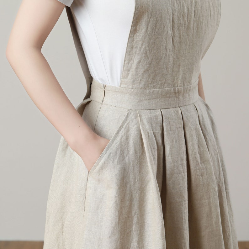 May include: A beige linen overall dress with a pleated skirt and pockets. The dress has a simple, casual style.
