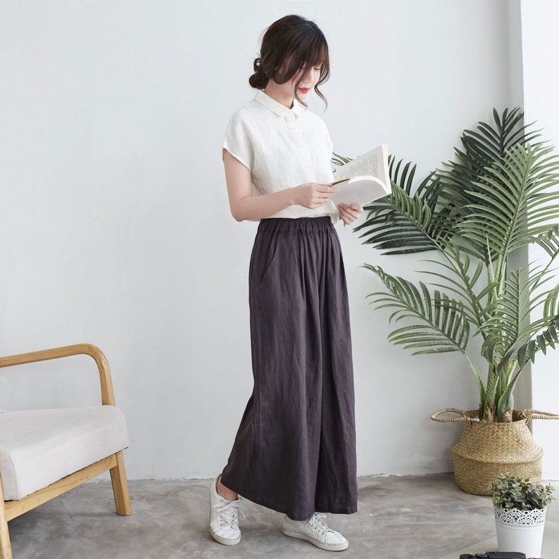 Puede incluir: Una mujer con una camisa de lino blanca y pantalones anchos de color gris oscuro. Est&aacute; sosteniendo un libro y mir&aacute;ndolo.