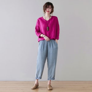May include: A woman wearing a magenta V-neck long-sleeve shirt and light blue denim pants. The pants have an elastic waistband and are cuffed at the ankles. She is also wearing a necklace and brown shoes.