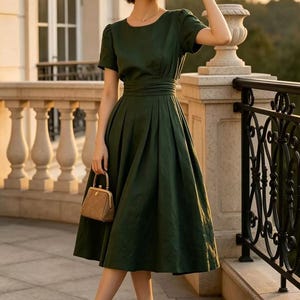 May include: A dark green midi dress with short sleeves, a round neckline, and a pleated skirt. The dress features a wide, gathered waistband. A tan straw hat, a small tan handbag with a gold frame, and tan pumps with black toes complete the look.