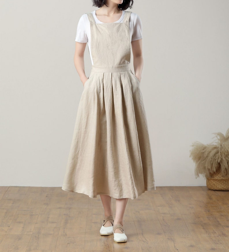 May include: A beige linen overall dress with a pleated skirt and thin straps. The dress is midi length and has a relaxed fit.