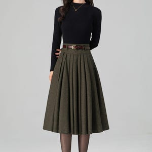 May include: A woman's outfit featuring a black long-sleeved top and a pleated olive green skirt. A brown belt cinches the waist. The outfit is completed with brown heeled boots and sheer black tights. The overall style is classic and elegant.