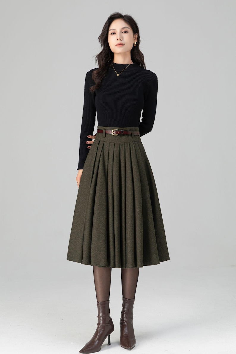 May include: A woman's outfit featuring a black long-sleeved top and a pleated olive green skirt. A brown belt cinches the waist. The outfit is completed with brown heeled boots and sheer black tights. The overall style is classic and elegant.