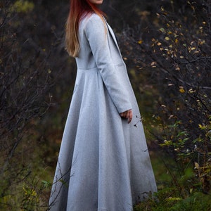 May include: A light gray, long coat with a flared skirt and a tailored fit. The coat has a classic design with a lapel collar and long sleeves. The person in the image is wearing the coat in a natural outdoor setting.