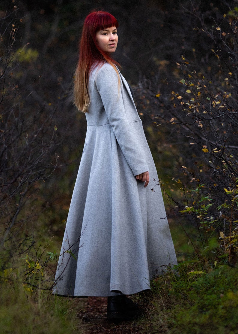 May include: A light gray, long coat with a flared skirt and a tailored fit. The coat has a classic design with a lapel collar and long sleeves. The person in the image is wearing the coat in a natural outdoor setting.
