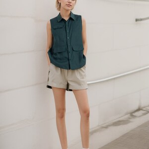 May include: A woman wearing a dark green sleeveless button-up shirt and beige shorts. She is standing in front of a white wall with silver pipes. She is wearing white sneakers.