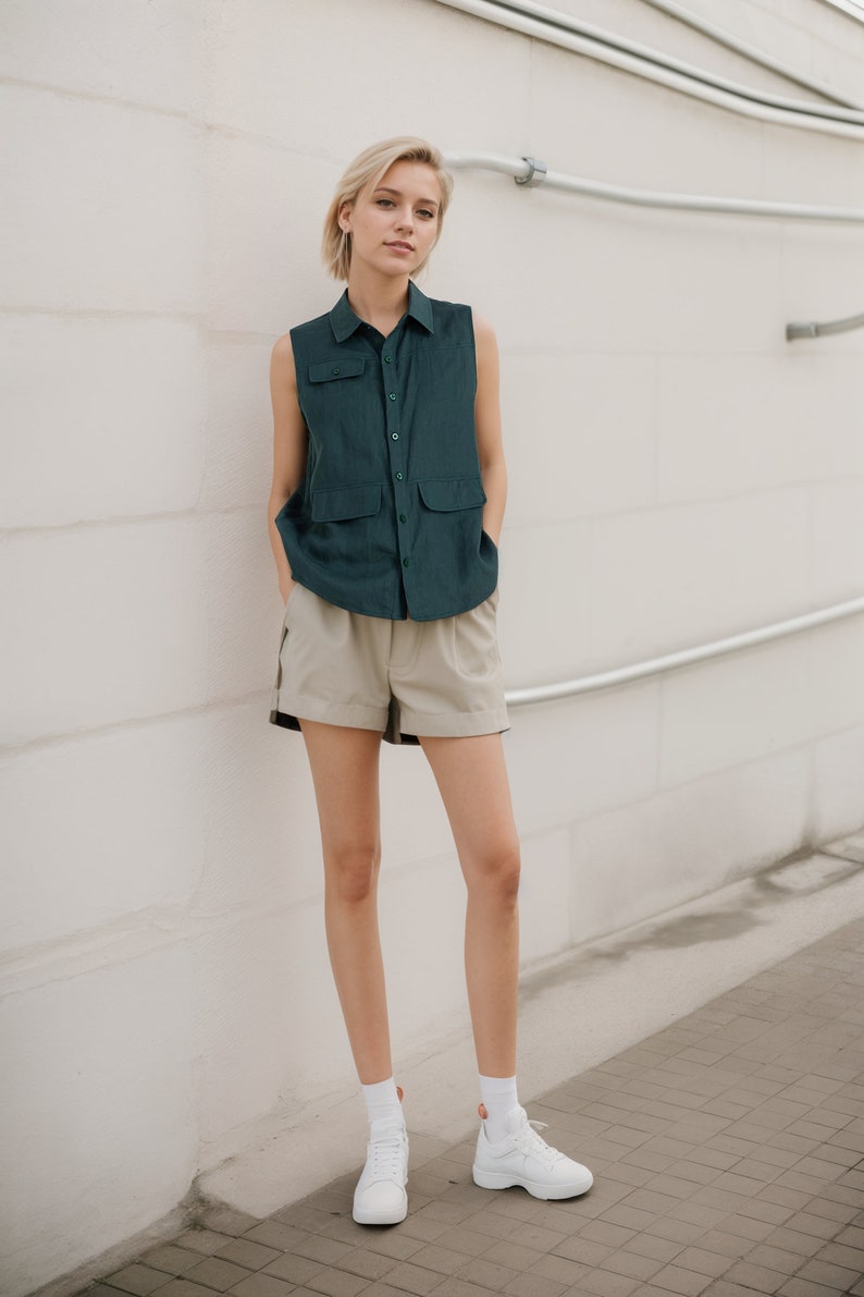 May include: A woman wearing a dark green sleeveless button-up shirt and beige shorts. She is standing in front of a white wall with silver pipes. She is wearing white sneakers.