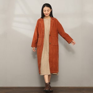 May include: A long, rust-colored corduroy coat with a button closure and two patch pockets. The coat is worn over a beige dress.