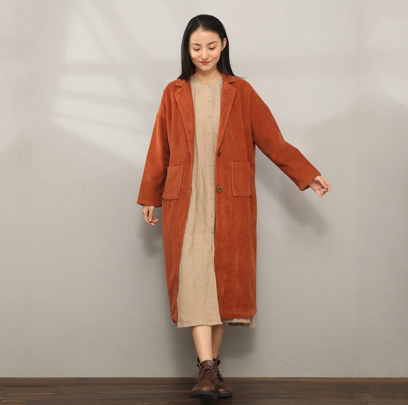 May include: A long, rust-colored corduroy coat with a button closure and two patch pockets. The coat is worn over a beige dress.