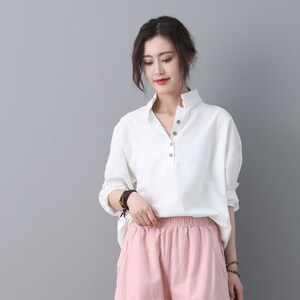 May include: A white linen shirt with a v-neck and button closure. The shirt has long sleeves and a relaxed fit. The model is wearing the shirt with pink linen pants.