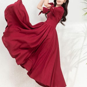 May include: A woman wearing a long, burgundy-colored dress with a tie detail at the sleeves. The dress has a fitted bodice and a flowing skirt. She is wearing black high heels.