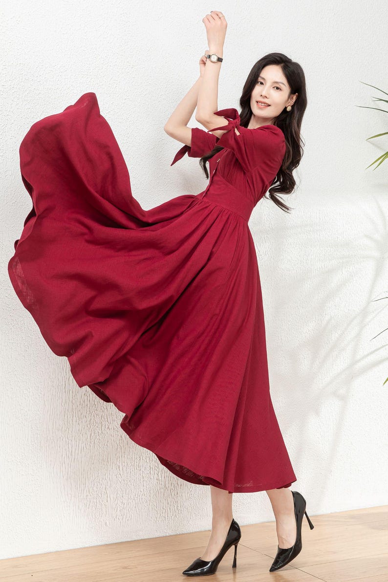 May include: A woman wearing a long, burgundy-colored dress with a tie detail at the sleeves. The dress has a fitted bodice and a flowing skirt. She is wearing black high heels.